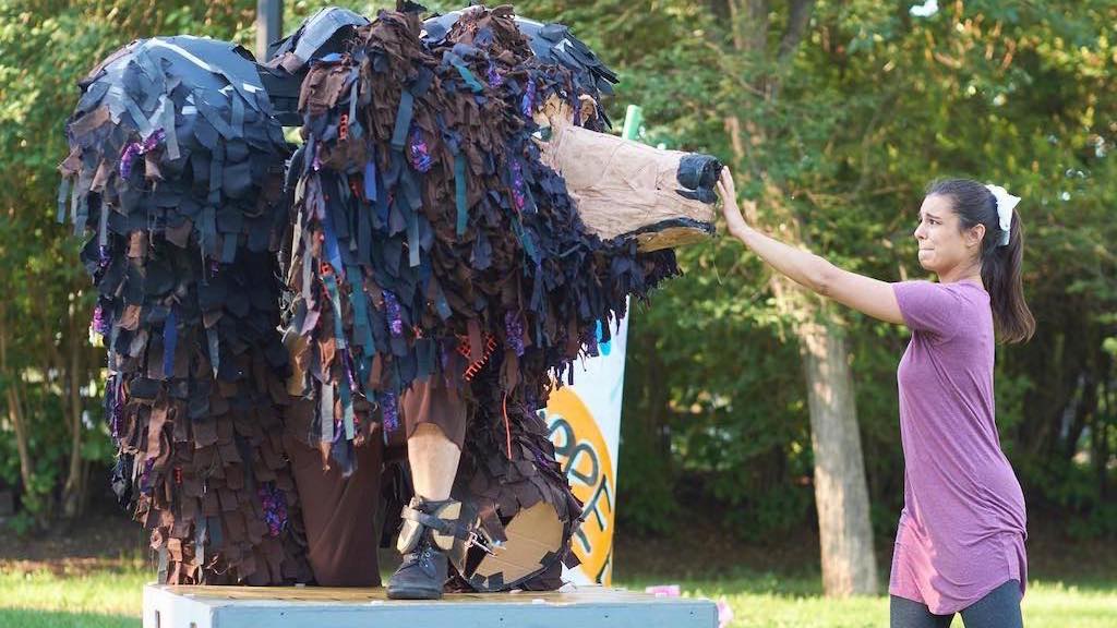 A woman in a purple shirt high-fives a large, person-operated bear puppet made of layered black and brown fabric strips with a paper-mch snout, standing on a platform in an outdoor park setting. Trees and a colorful sign are visible in the background.